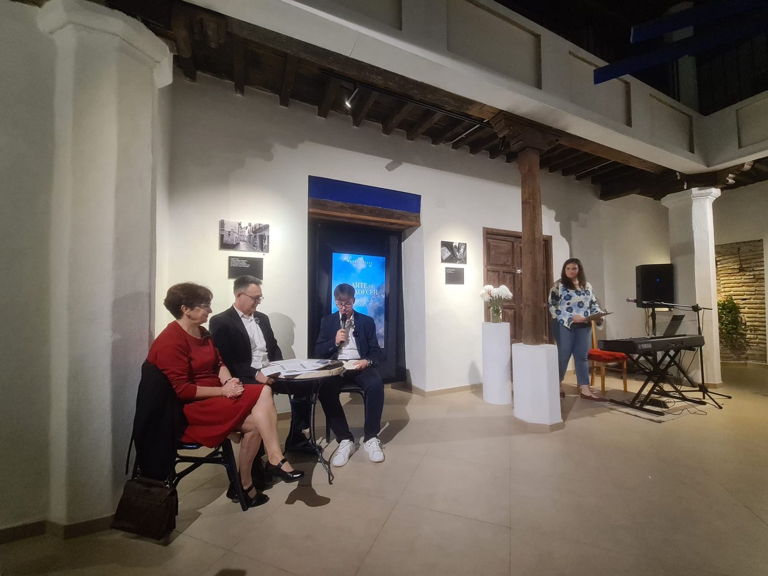 Mesa de la presentación junto a la pantalla con la portada del libro, una presentadora de pie y un teclado a la derecha