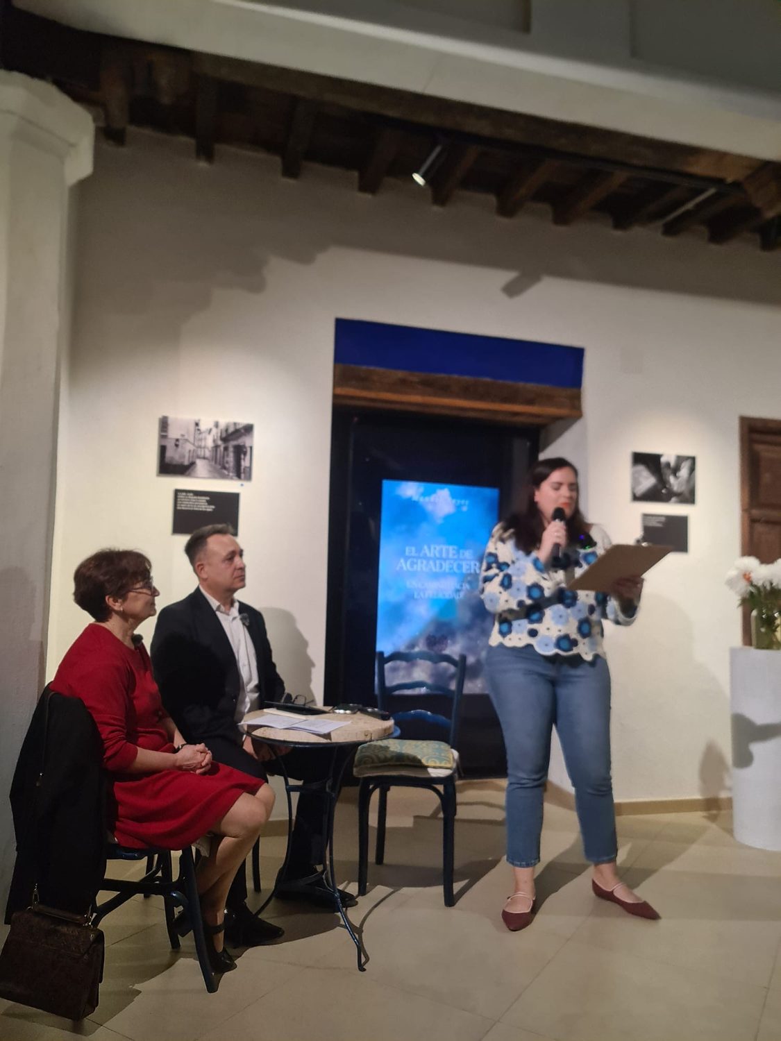 Una presentadora lee el programa del acto junto a la mesa, con la portada del libro de fondo