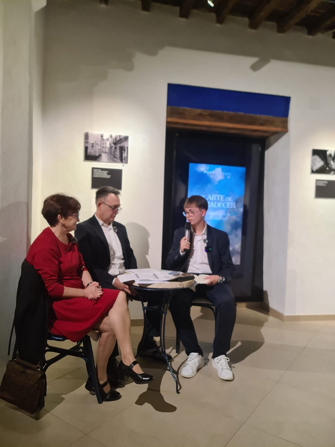 Tres personas conversan en la mesa de la presentación delante de la pantalla con la portada del libro